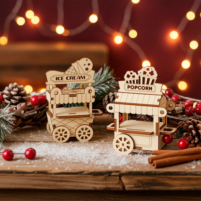 A pair of detailed laser cut wooden plywood miniature street carts—one for "ICE CREAM" and one for "POPCORN"—set on a wooden surface dusted with faux snow, surrounded by pine cones and red berries, with warm string lights in the background.