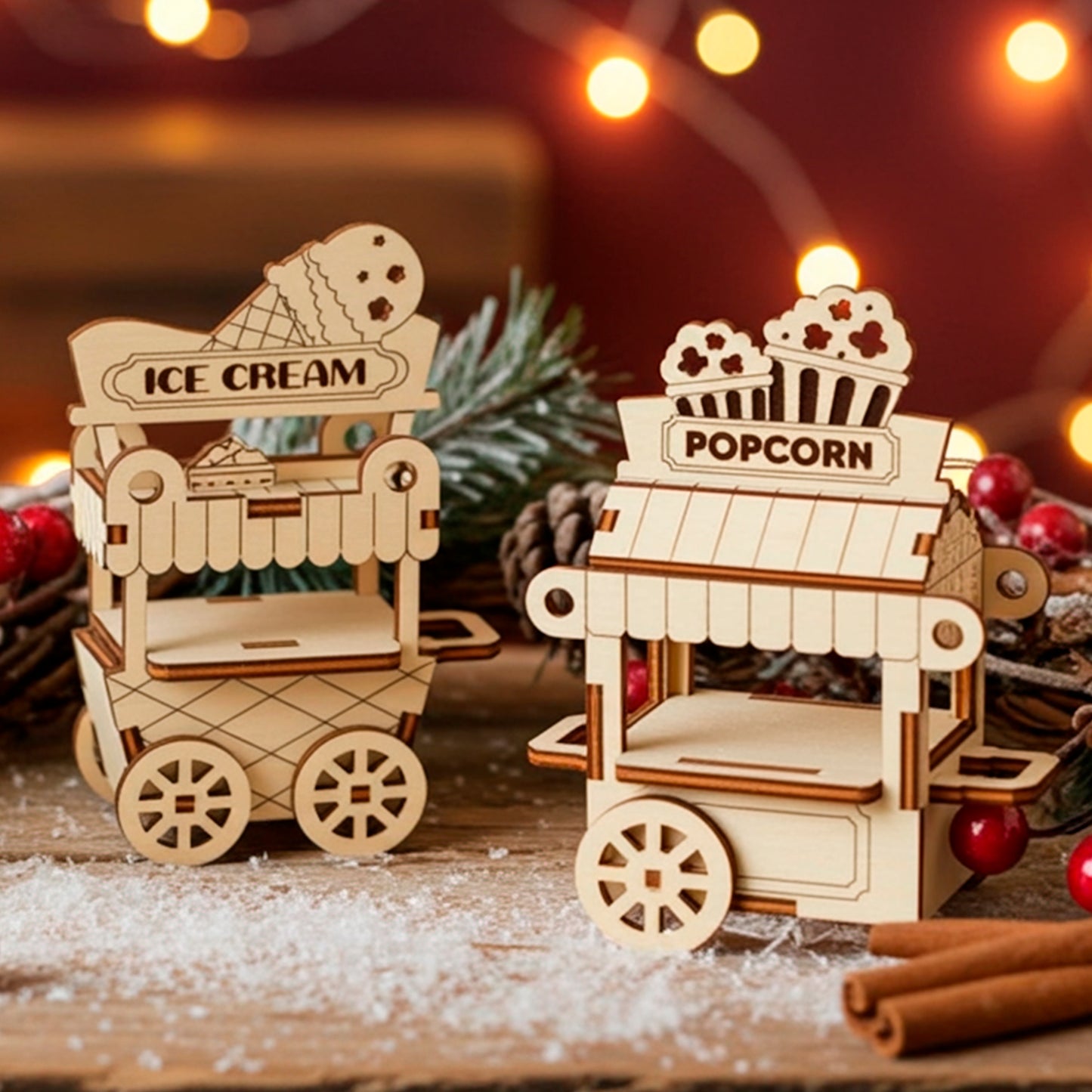 A pair of detailed laser cut wooden plywood miniature street carts—one for "ICE CREAM" and one for "POPCORN"—set on a wooden surface dusted with faux snow, surrounded by pine cones and red berries, with warm string lights in the background.