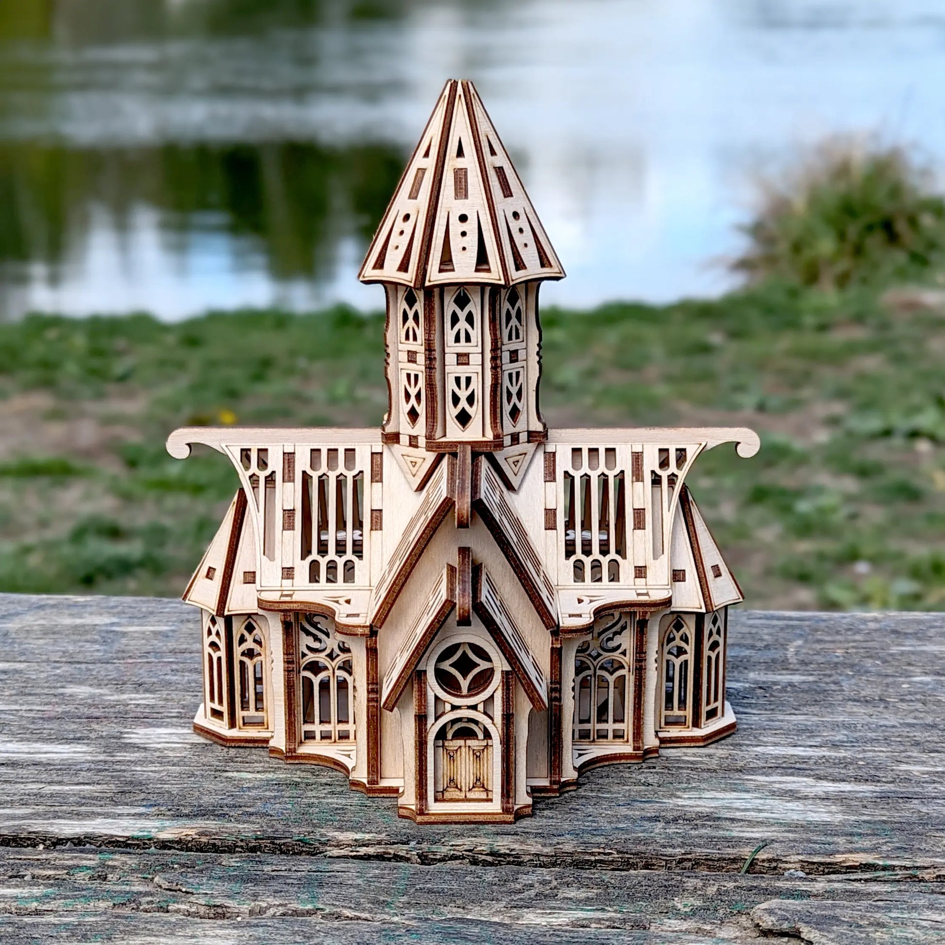 Sharp straight shot of a mini laser cut plywood gothic cathedral standing on an old wooden bench near a blurry lake.