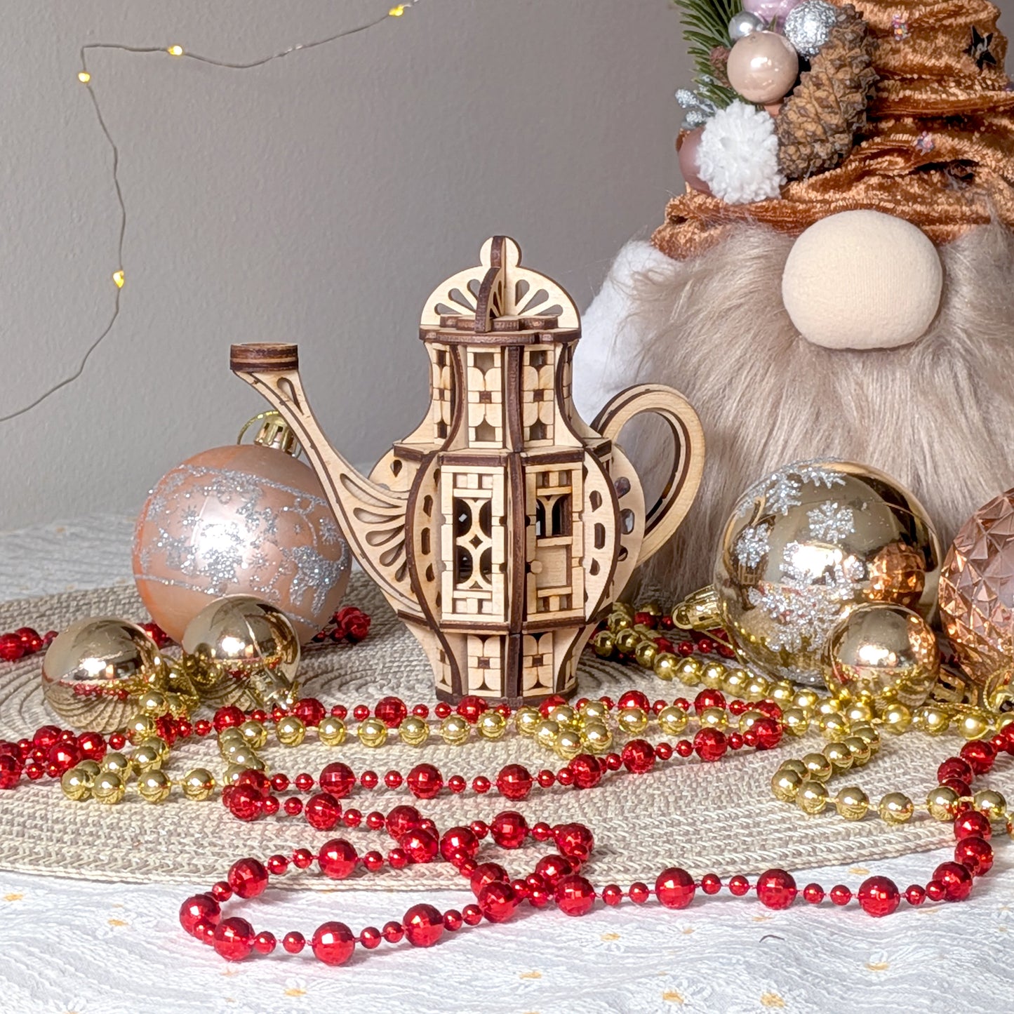 Laser cut wooden teapot fairy house displayed in a festive scene with Christmas ornaments, golden and red bead garlands, and soft lighting.