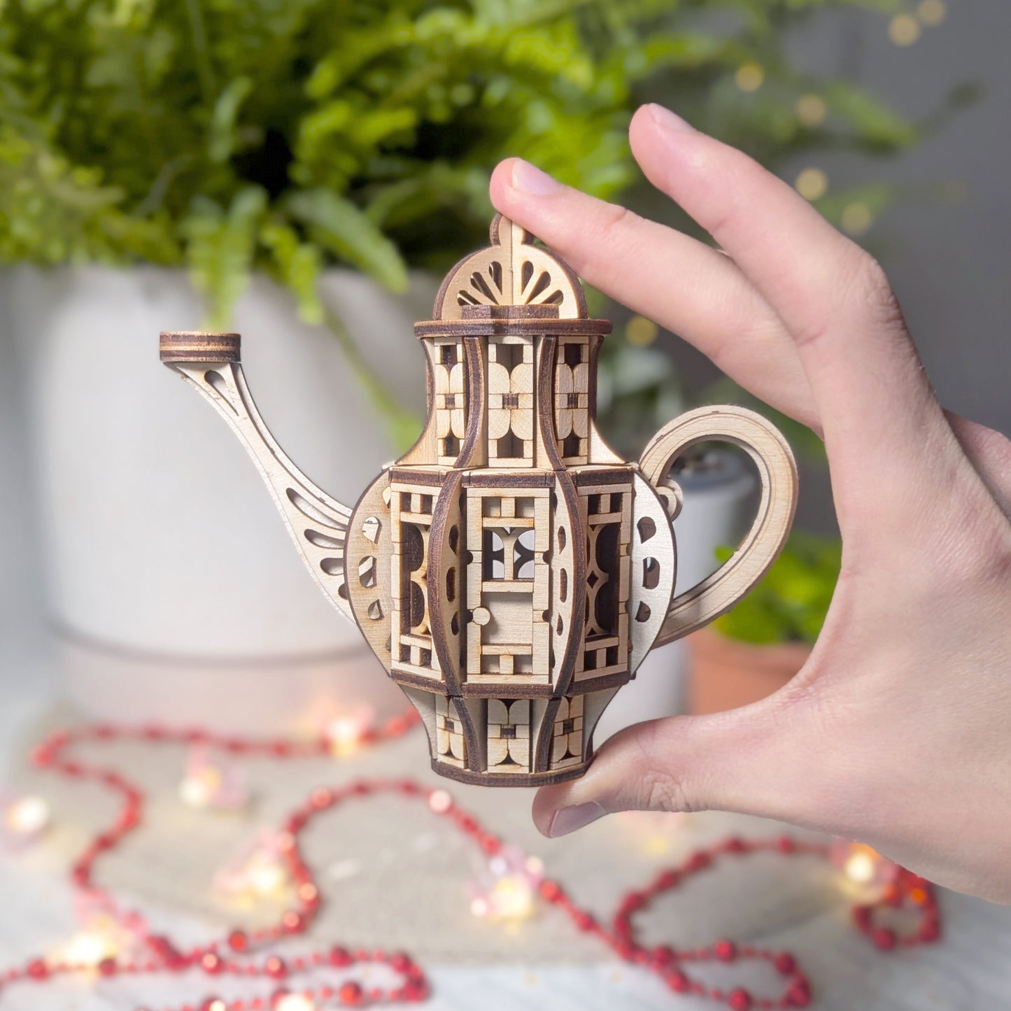 A hand holding a small laser cut wooden teapot fairy house, showing its compact size next to a plant and decorative lights.