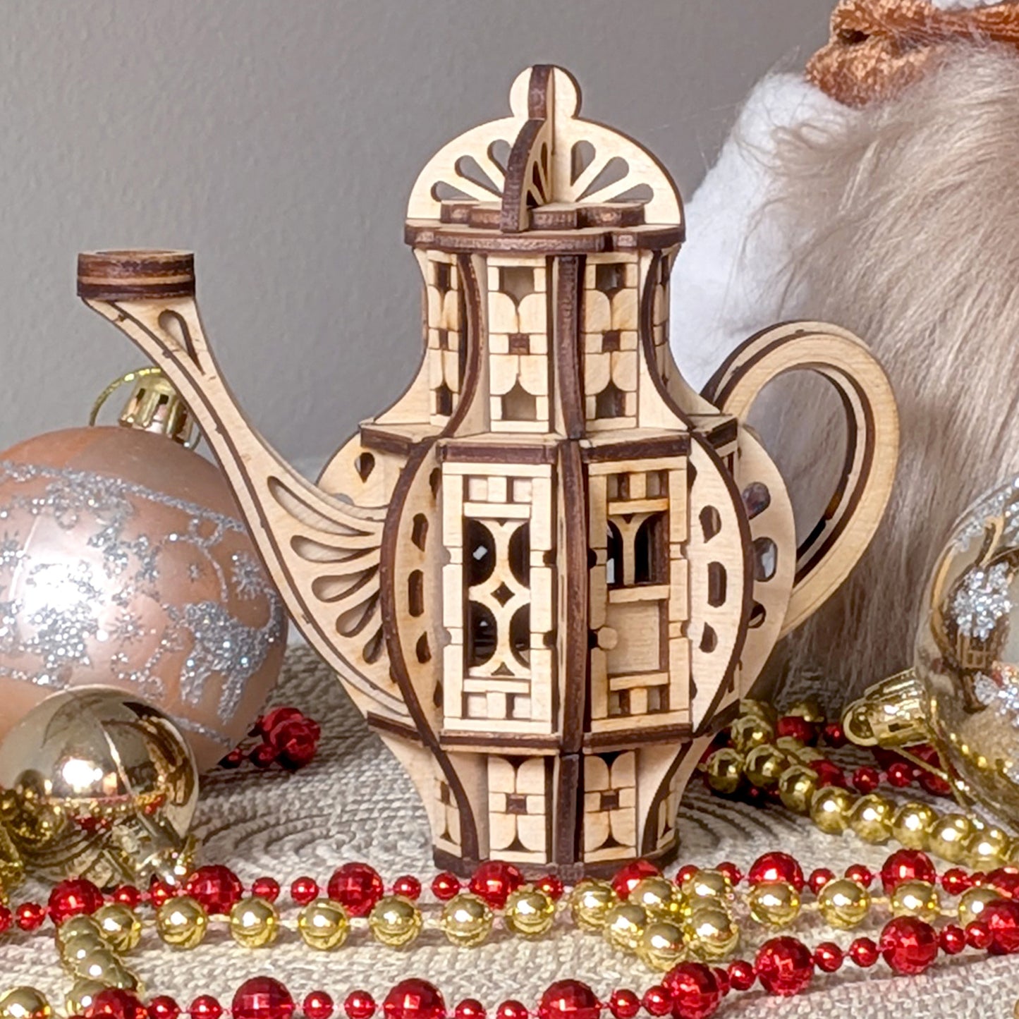 Laser cut wooden teapot-shaped fairy house model with decorative cutout panels and curved handle, displayed as holiday decor among ornaments and beaded garlands