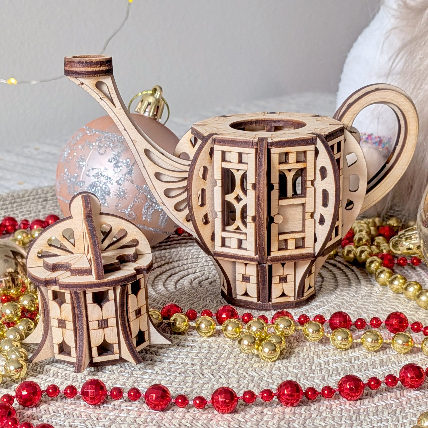 Laser cut wooden teapot fairy house with removable lid, placed on a woven mat alongside potted plants and decorative pink lights