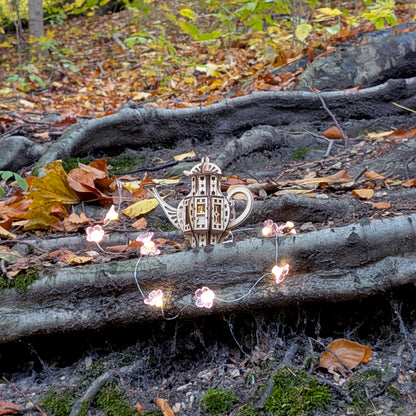 Laser cut wooden teapot fairy house set among tree roots in a forest setting, illuminated by small glowing flower-shaped lights