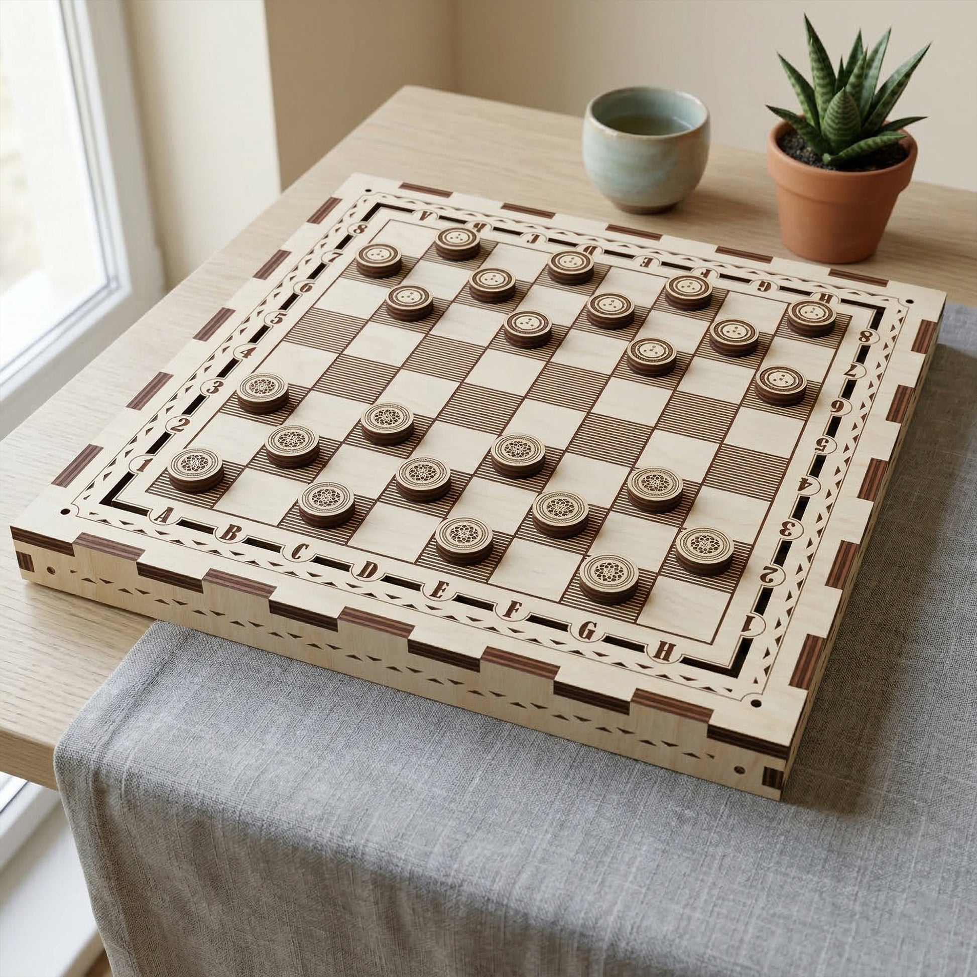 Laser cut plywood checkers set on a matching wooden board. Round checkers feature detailed engraved patterns. The board has decorative borders and coordinate letters. Styled on a table with a grey linen runner and a small potted succulent.