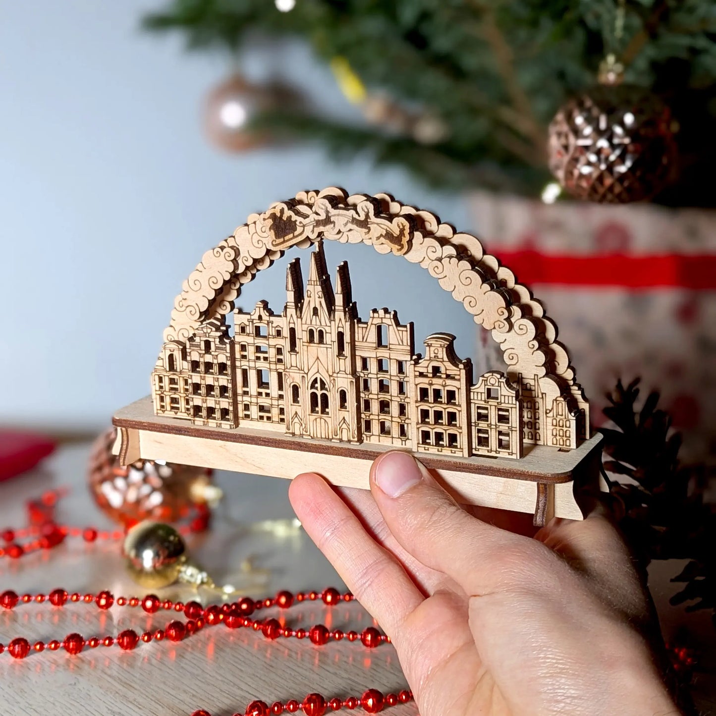 A person's hand holding a miniature 3D Christmas town arch to show scale, beautifully crafted from premium laser cut plywood.
