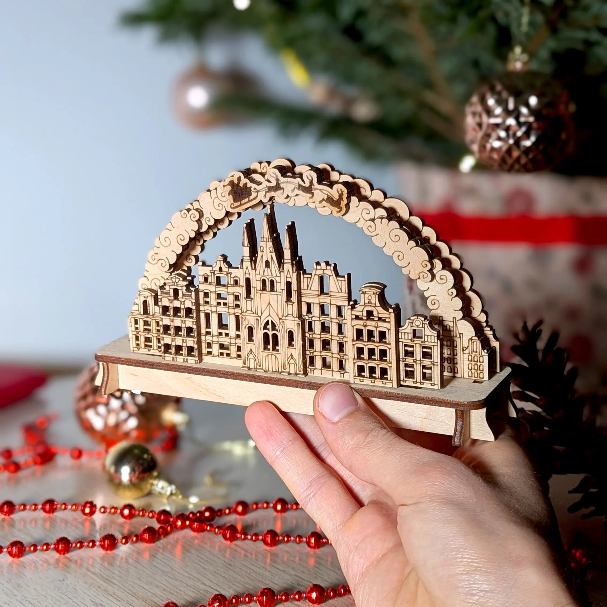 A person's hand holding a miniature 3D Christmas town arch to show scale, beautifully crafted from premium laser cut plywood.