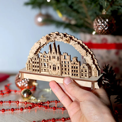 A person's hand holding a miniature 3D Christmas town arch to show scale, beautifully crafted from premium laser cut plywood.