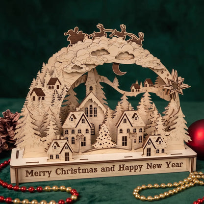 A laser-cut wooden Christmas village tabletop decoration with a snowy hillside village, pine trees, a crescent moon, stars, and Santa in a sleigh, displayed on a green fabric background with red and gold beads and a red ornament nearby.