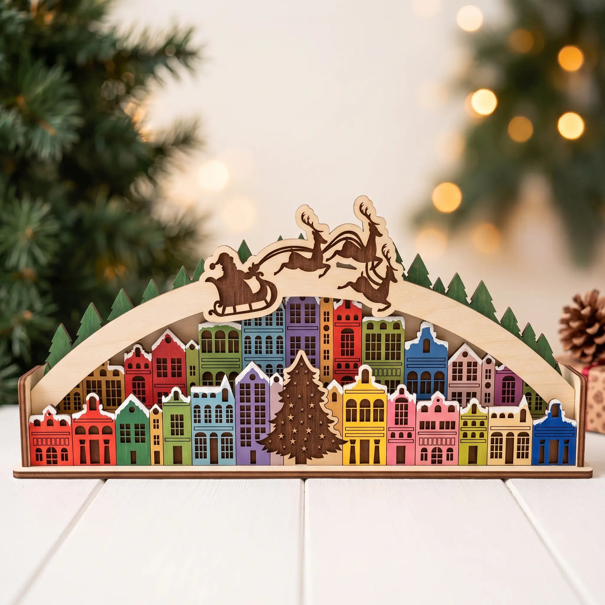 Painted laser cut plywood Christmas town arch diorama on white wooden tabletop in front of decorated tree with warm bokeh.