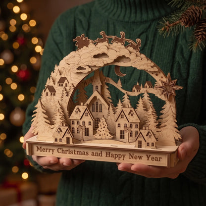 A person in a green sweater holds a laser-cut wooden Christmas village scene with a base that reads 'Merry Christmas and Happy New Year.' The background shows a softly blurred Christmas tree with warm lights.