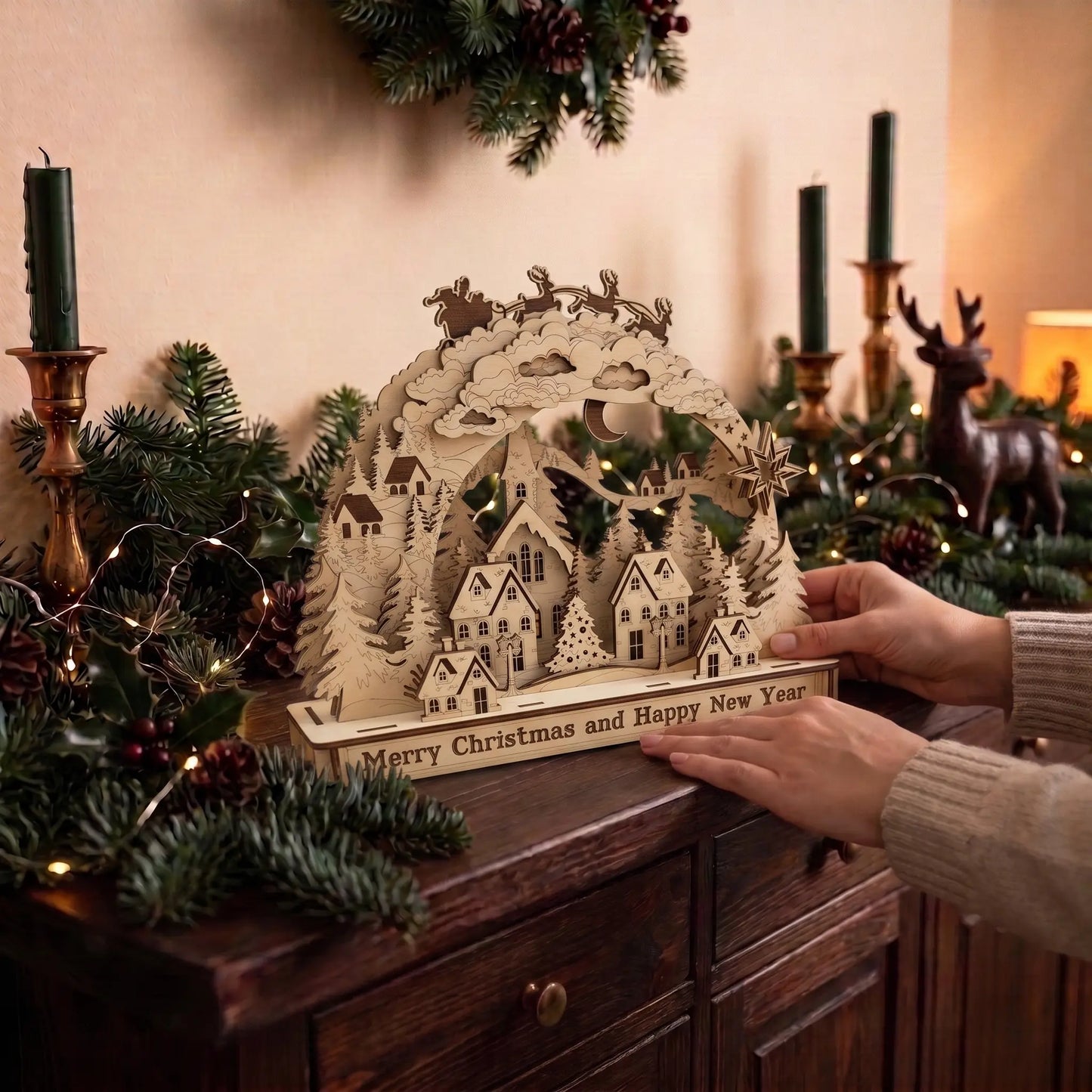 A wooden laser-cut Christmas village tabletop display with houses, trees, a crescent moon, and a deer silhouette, held by hands on a dark wooden cabinet surrounded by greenery, candles, and warm holiday decor.