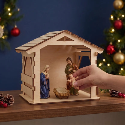 Hand places baby Jesus into manger inside laser cut plywood nativity stable beside Mary and Joseph on wood shelf.