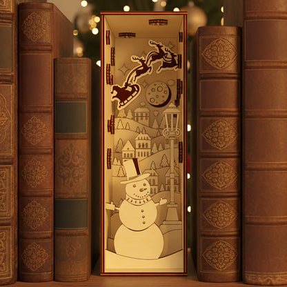 Laser cut wooden Christmas Book Nook featuring a cheerful snowman in the foreground, Santa's sleigh flying over a snowy village and glowing moon, displayed between vintage books against a warm Christmas tree lights background.
