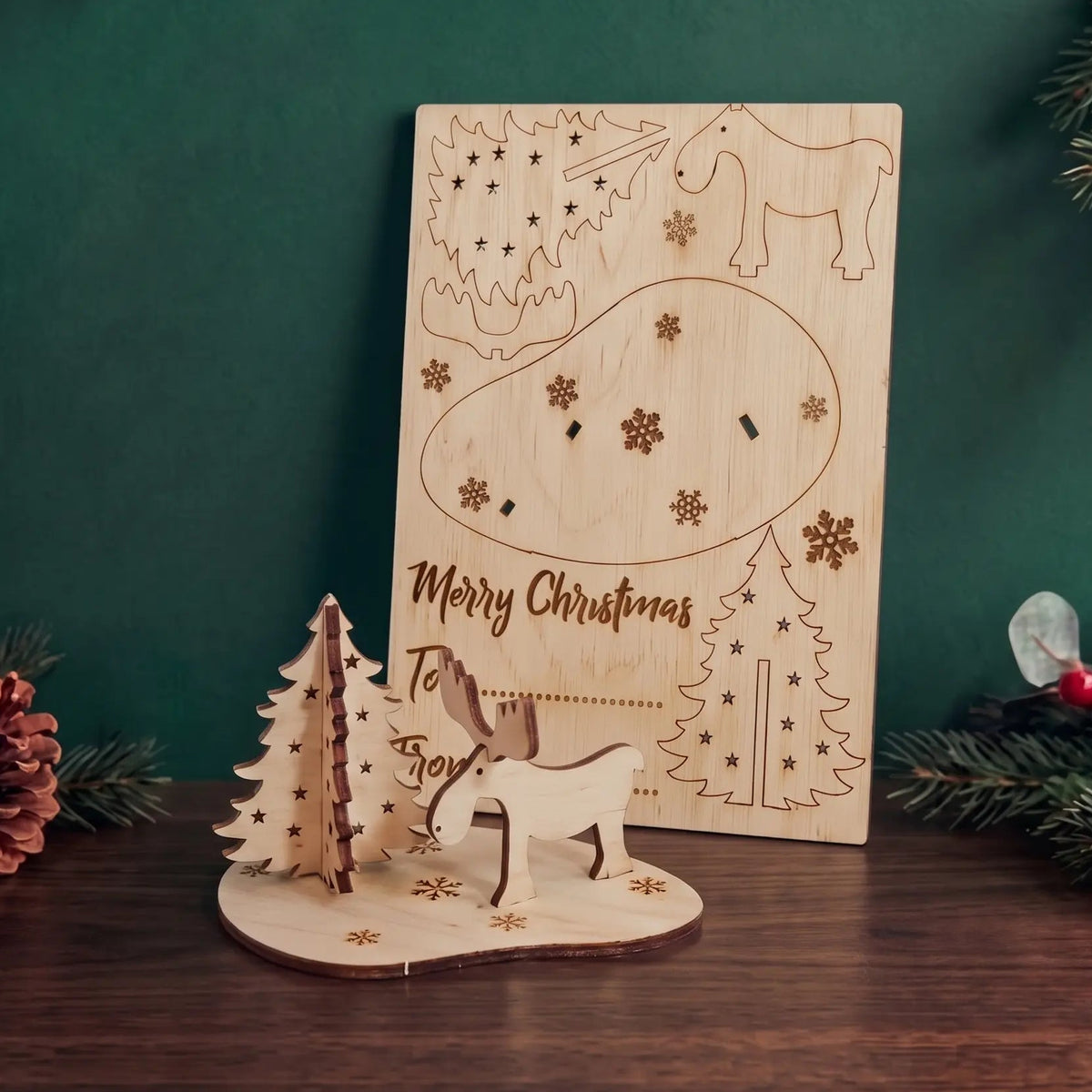Laser cut plywood holiday card with assembled miniature moose and tree set against dark green wall.