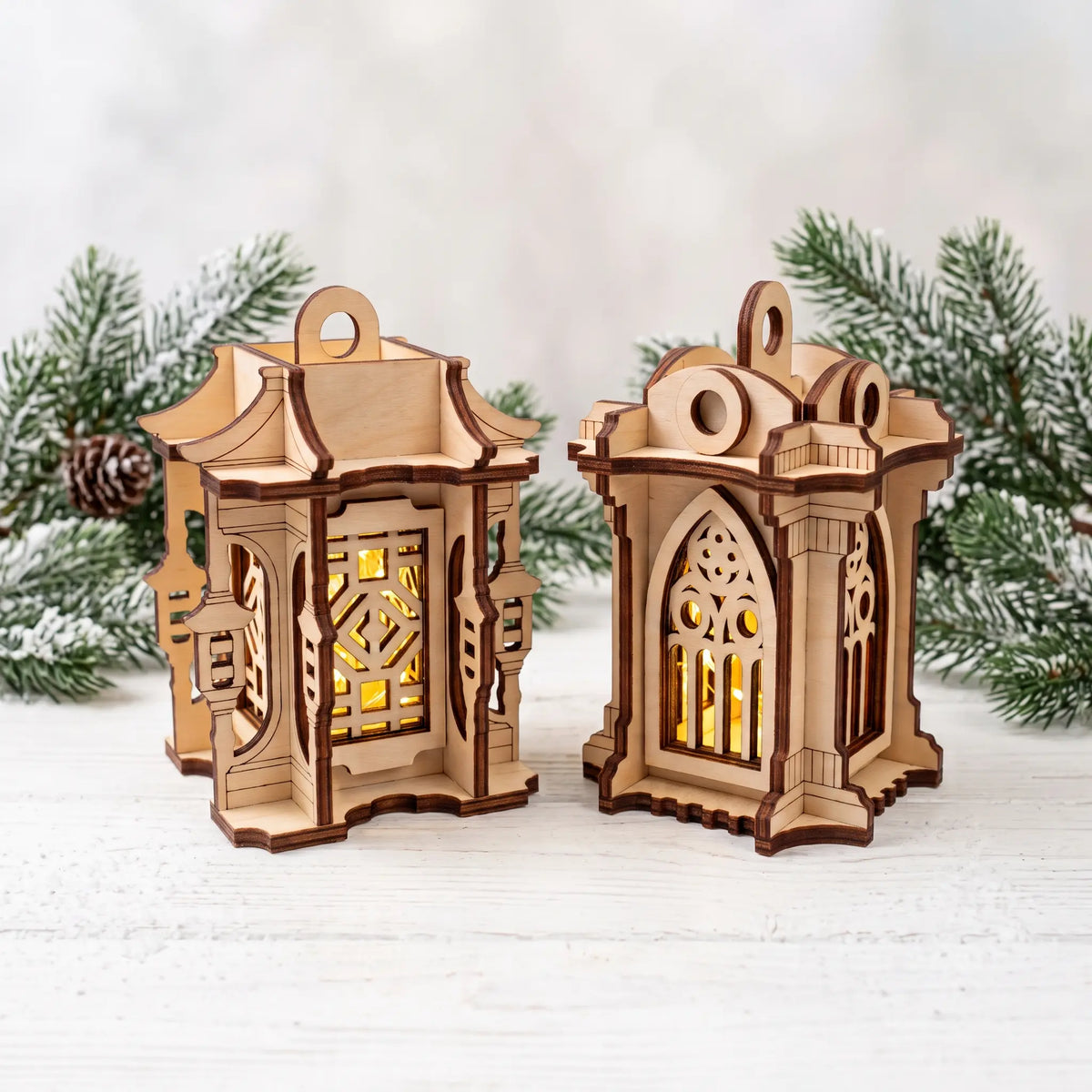 Two glowing laser cut plywood miniature lanterns with carved Asian and gothic arches on white table with pine branches.