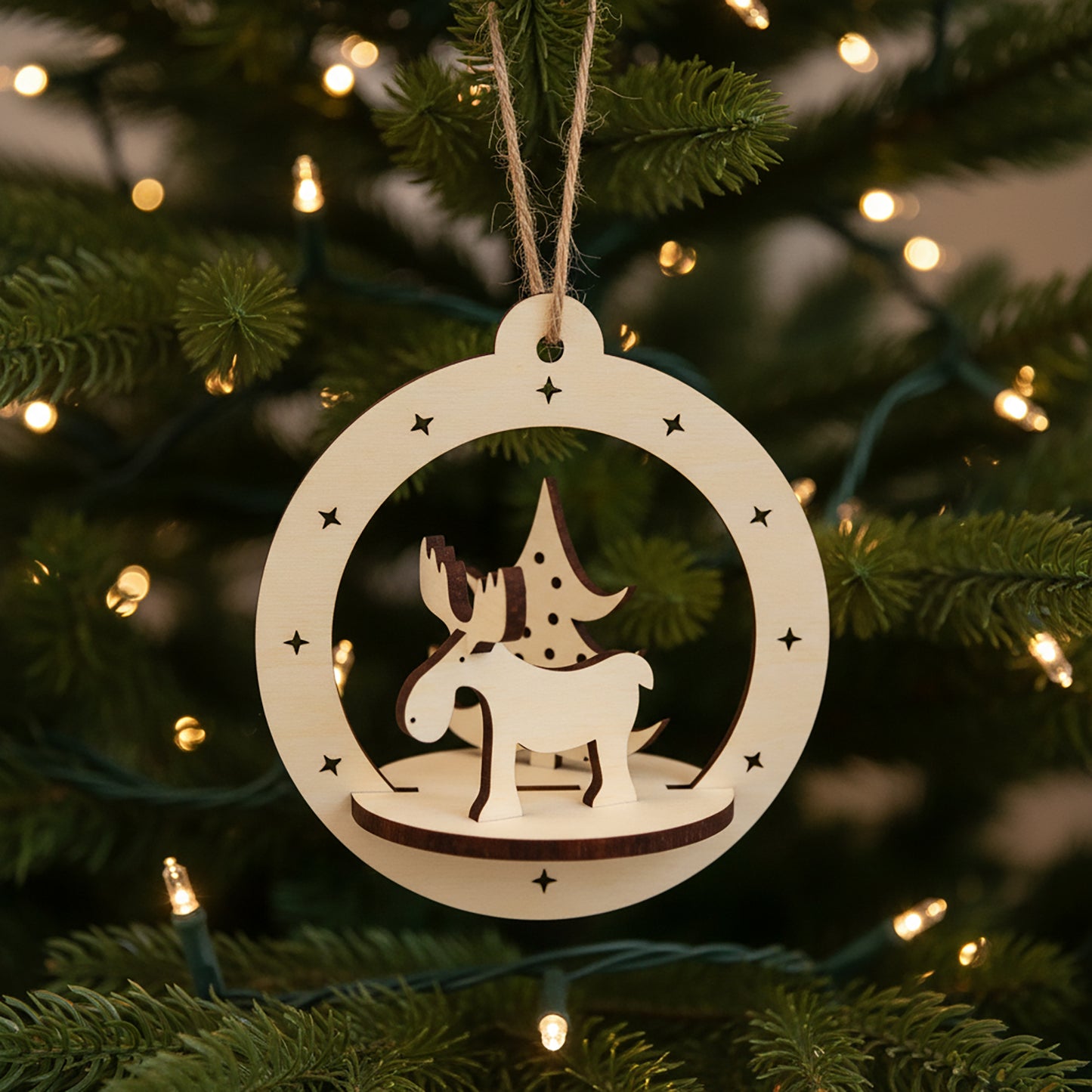 Intricate laser cut Christmas Deer Ornament featuring a cute reindeer with antlers and holiday tree in a hanging round frame with star accents, set against a warm lighted Christmas tree background.