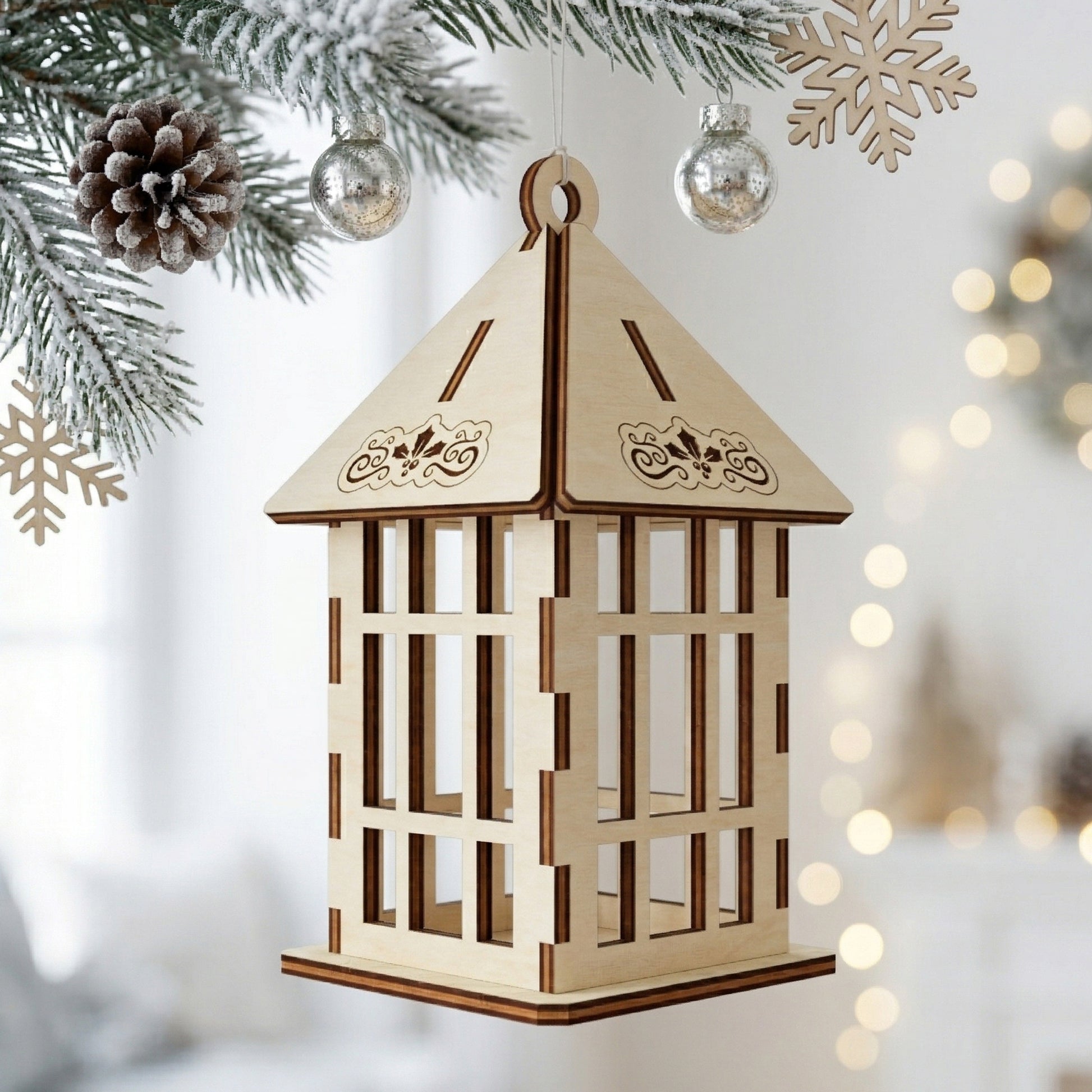 A single 3D laser-cut plywood lantern ornament with a classic lattice window design. The roof features engraved holly and leaf motifs. Displayed hanging on a snowy Christmas tree branch with silver baubles and pine cones in a bright, modern setting.