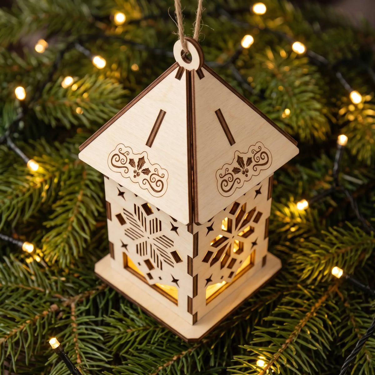 Top-down view of glowing laser cut plywood mini lantern sitting on fresh pine needles and fairy lights.