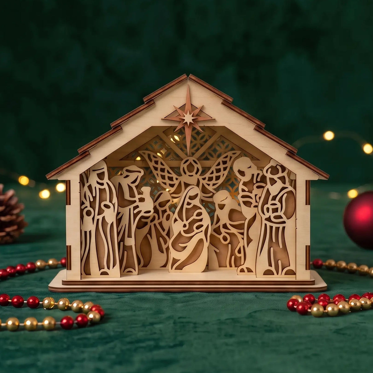 Illuminated laser cut plywood nativity crèche on green velvet with glowing fairy lights and red bead.