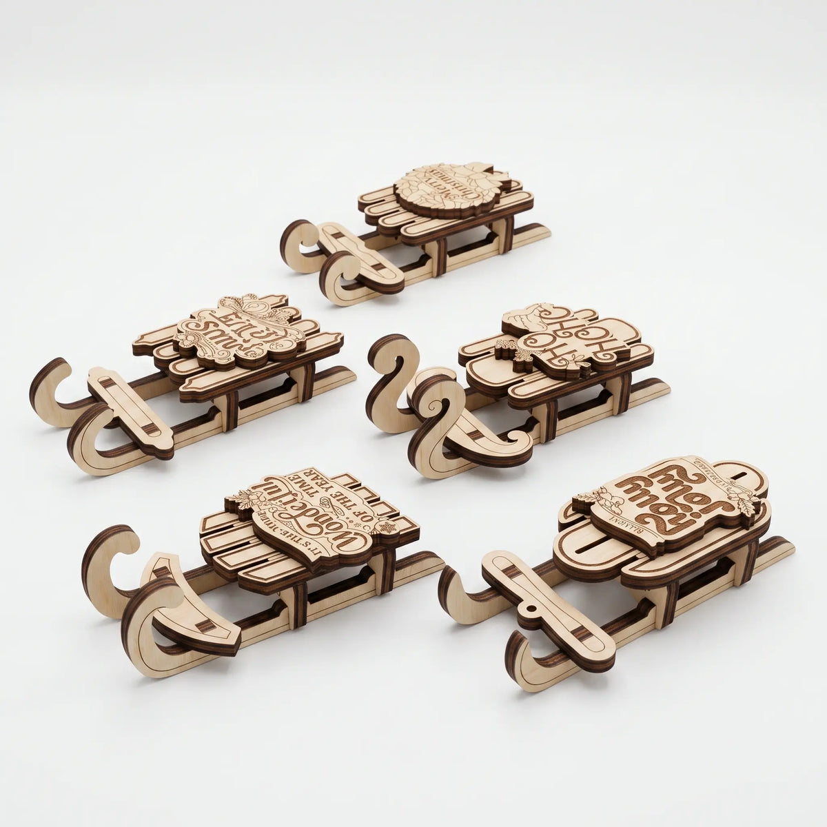 Five laser-cut wooden sledding ornaments in natural wood tones, arranged on a clean white background. Each ornament features a different holiday design