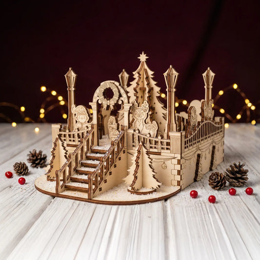 Staged photo of a laser cut plywood Christmas village on a white wood table with pinecones, faux snow, and fairy lights