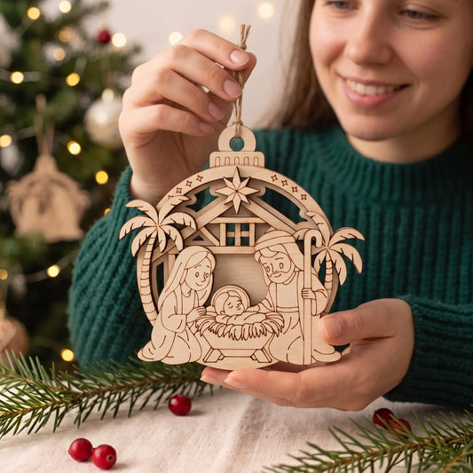 Woman holds layered laser cut plywood nativity bauble ornament over festive table with fir sprigs and lights.