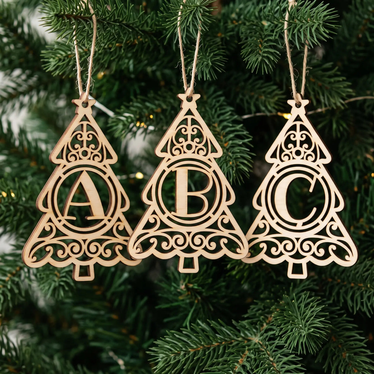 Three laser cut plywood Christmas tree ornaments with letters A, B, and C hanging on twine strings among green pine needles.