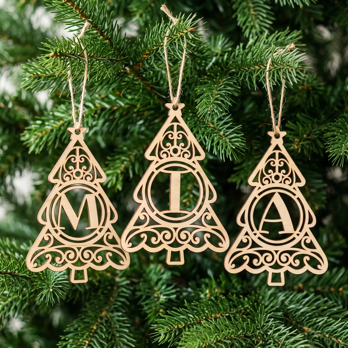 Three laser cut plywood monogram Christmas tree ornaments spelling MIA hanging by twine strings among evergreen branches.