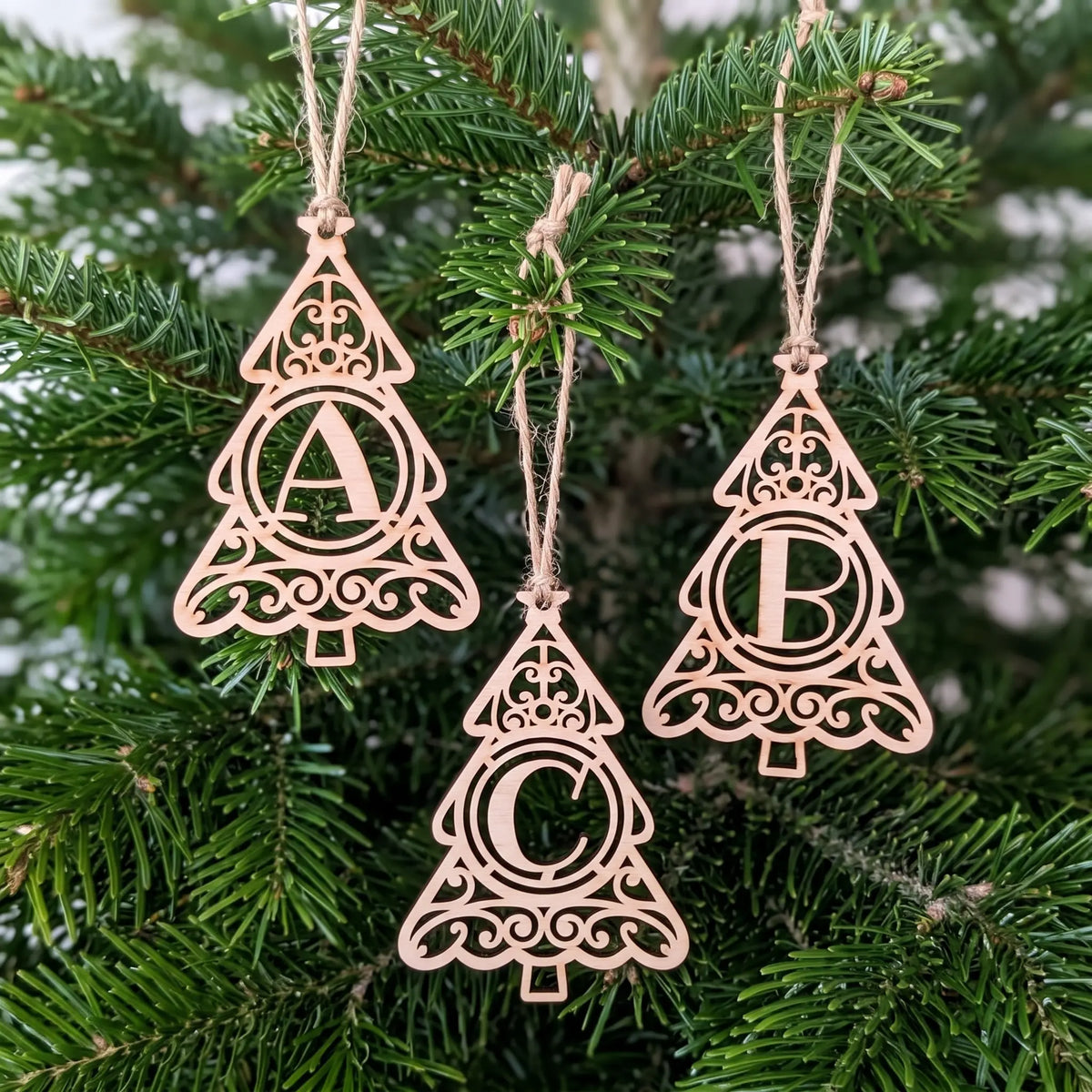 Staggered set of three laser cut plywood tree letter ornaments A, B, and C hanging by twine strings on festive pine tree.