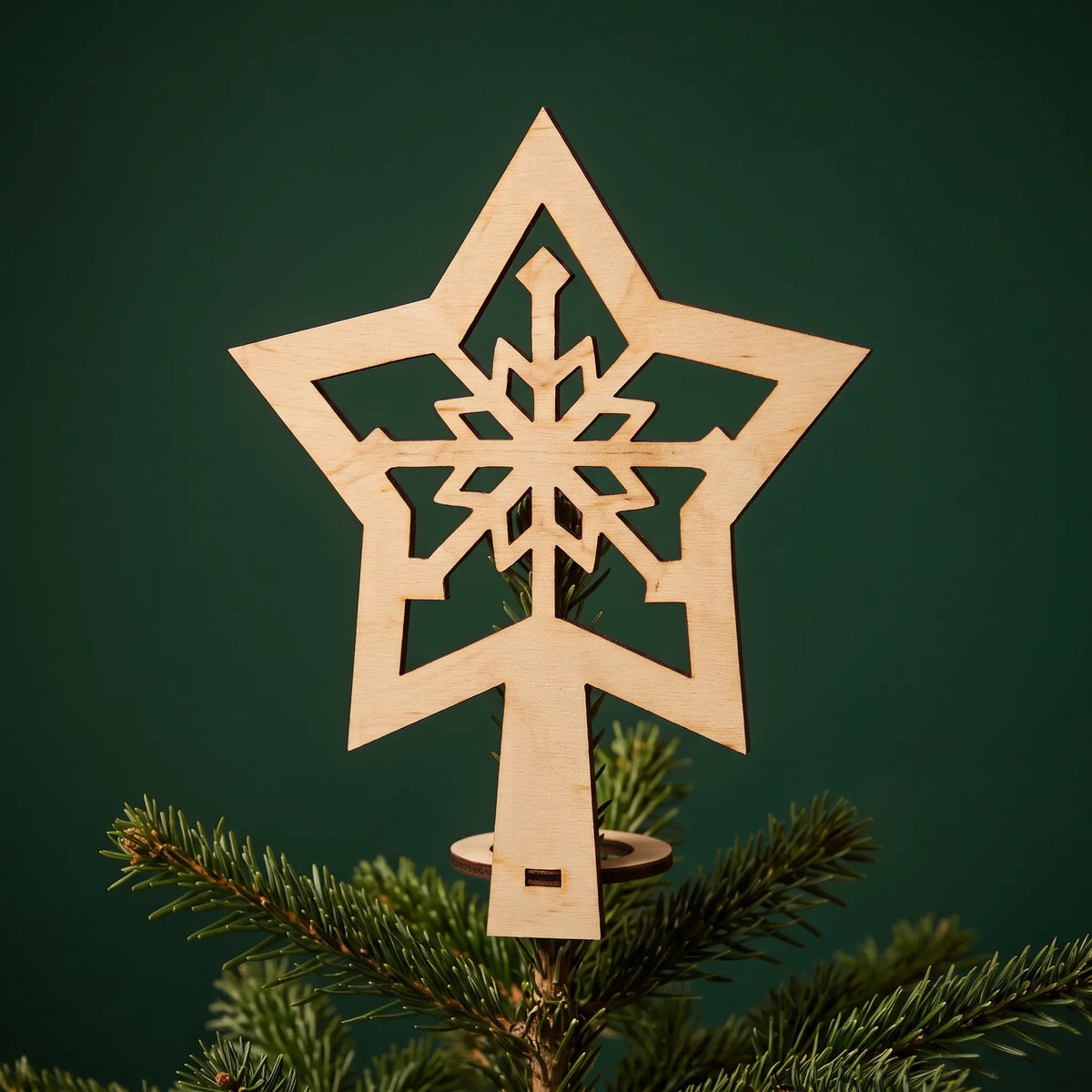 Natural laser cut plywood snowflake star tree topper mounted on Christmas tree branch against a dark green backdrop.