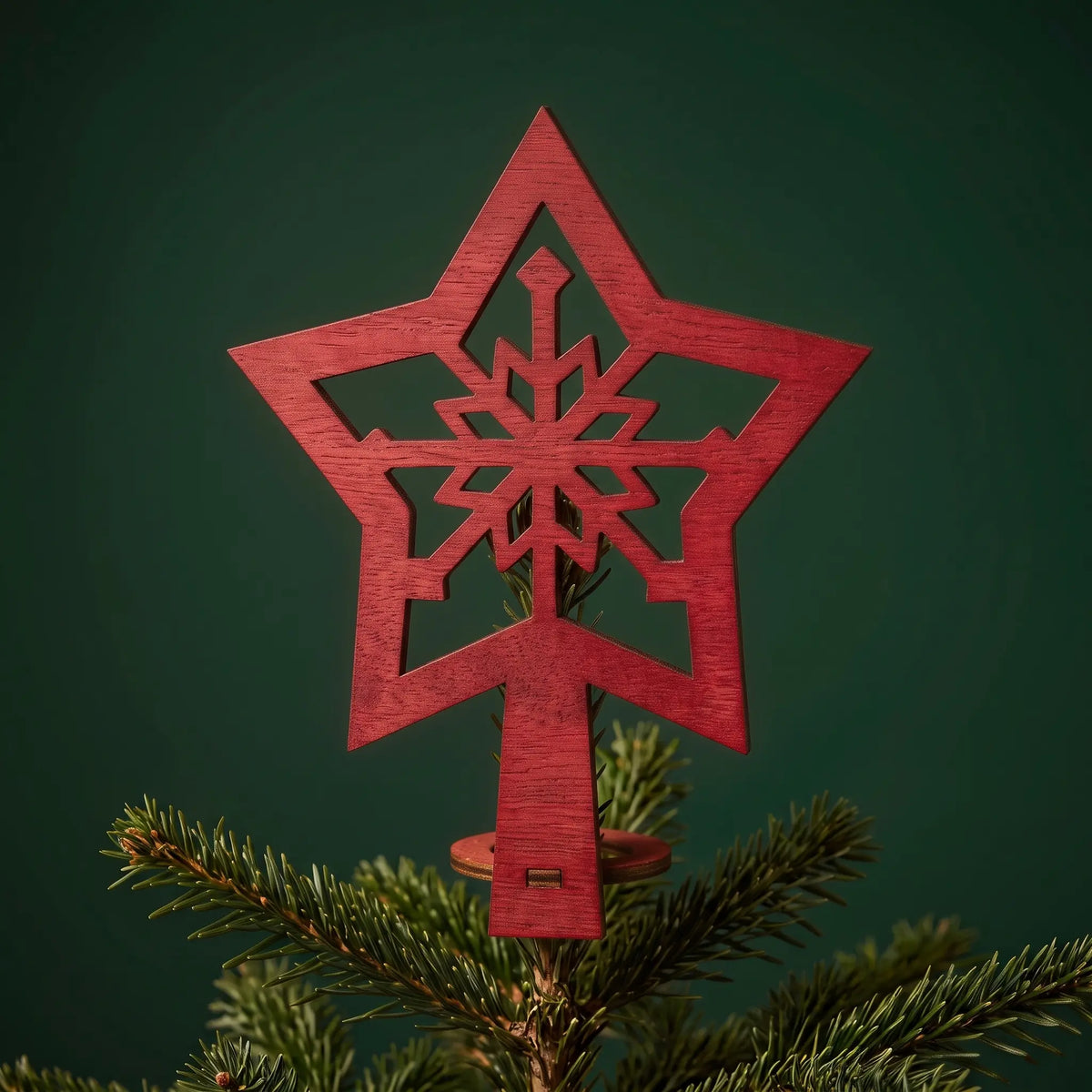 Red painted laser cut plywood snowflake star tree topper mounted on a green Christmas tree top against dark backdrop.