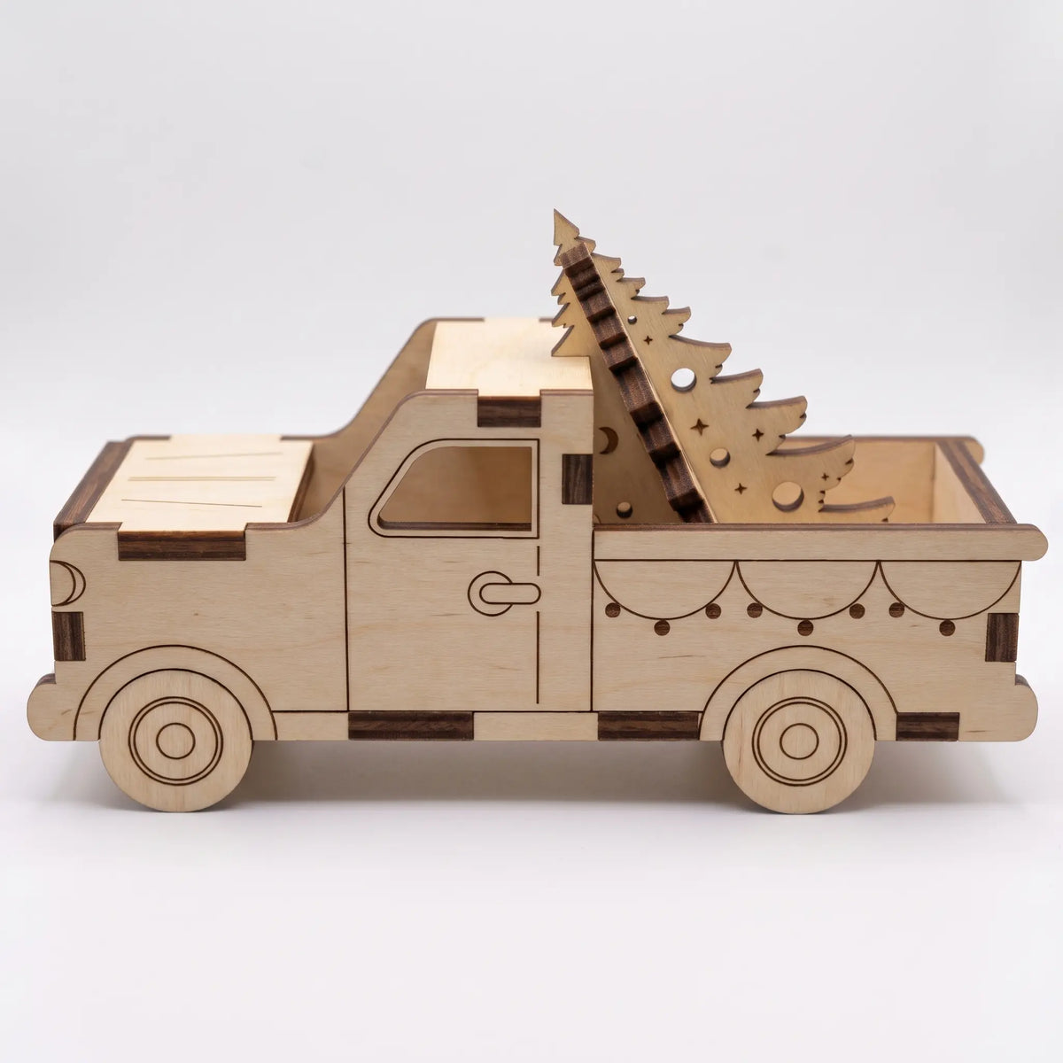 Side profile view of laser cut plywood holiday pickup truck model carrying cutout pine tree on plain white.