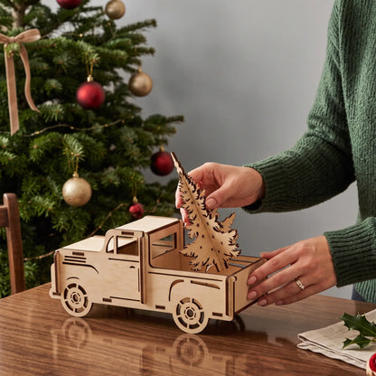 Woman positions laser cut plywood pine tree into truck bed on wooden tabletop beside decorated holiday tree.
