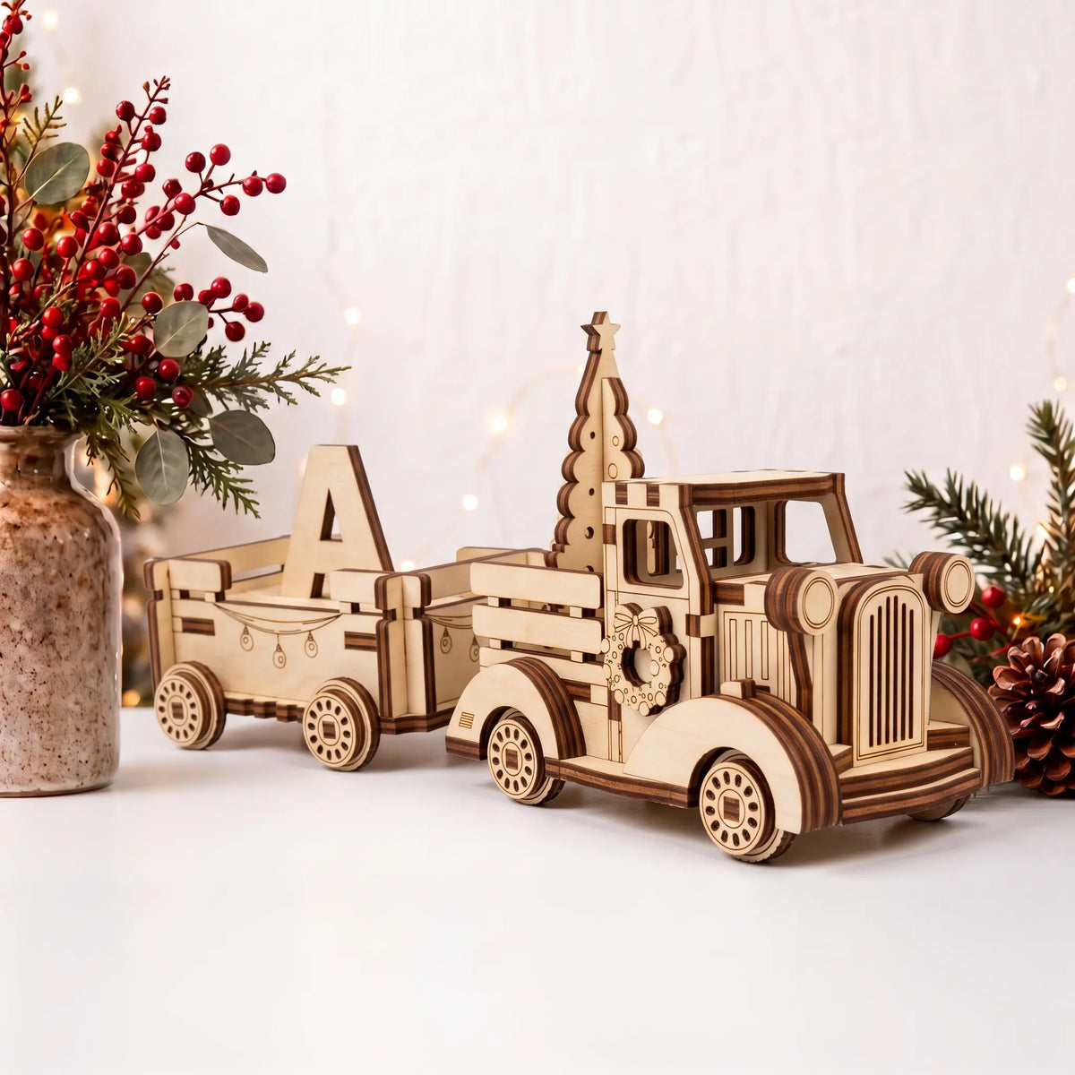 Laser cut plywood Christmas truck with tree and wagon hauling letter A on white table beside red holiday berries vase.