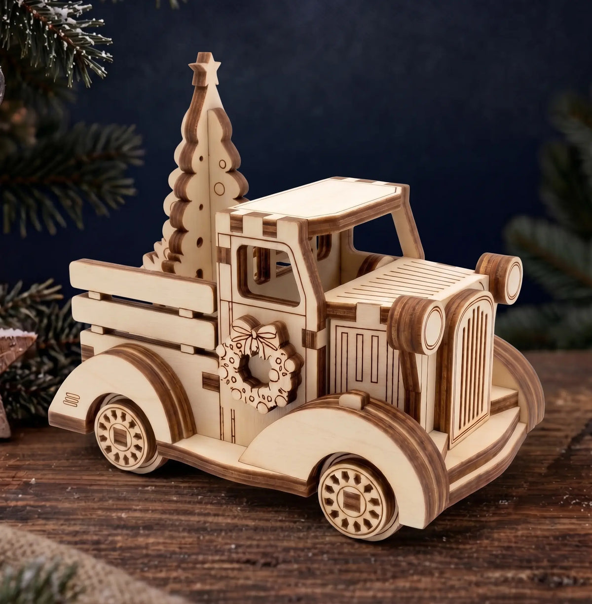 A wooden laser-cut Christmas truck ornament with a small Christmas tree in the bed, displayed on a rustic wooden surface with dark blue background and pine branches.