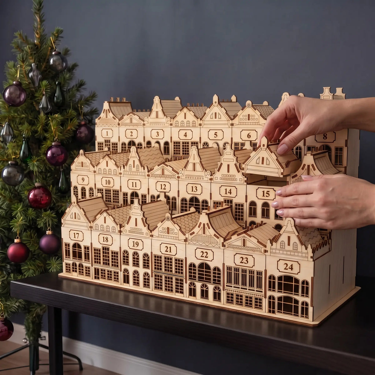 Hands remove small house drawer from tiered laser cut plywood Christmas village advent calendar beside holiday tree