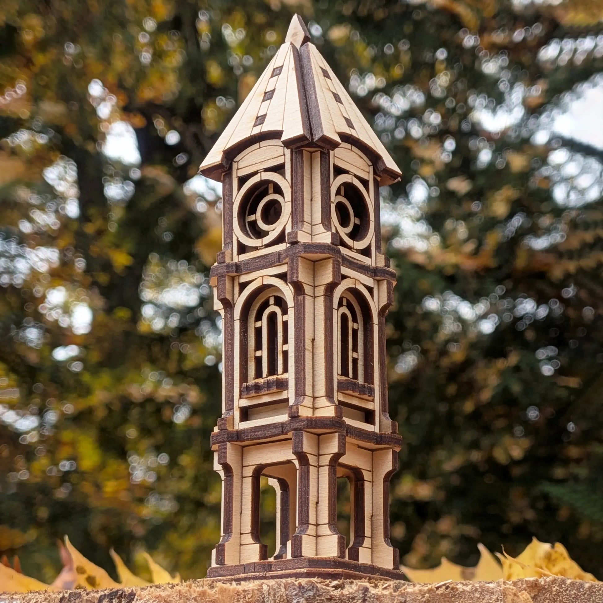 Low-angle close up of a miniature architectural clock tower model crafted from laser cut plywood against autumn trees bokeh