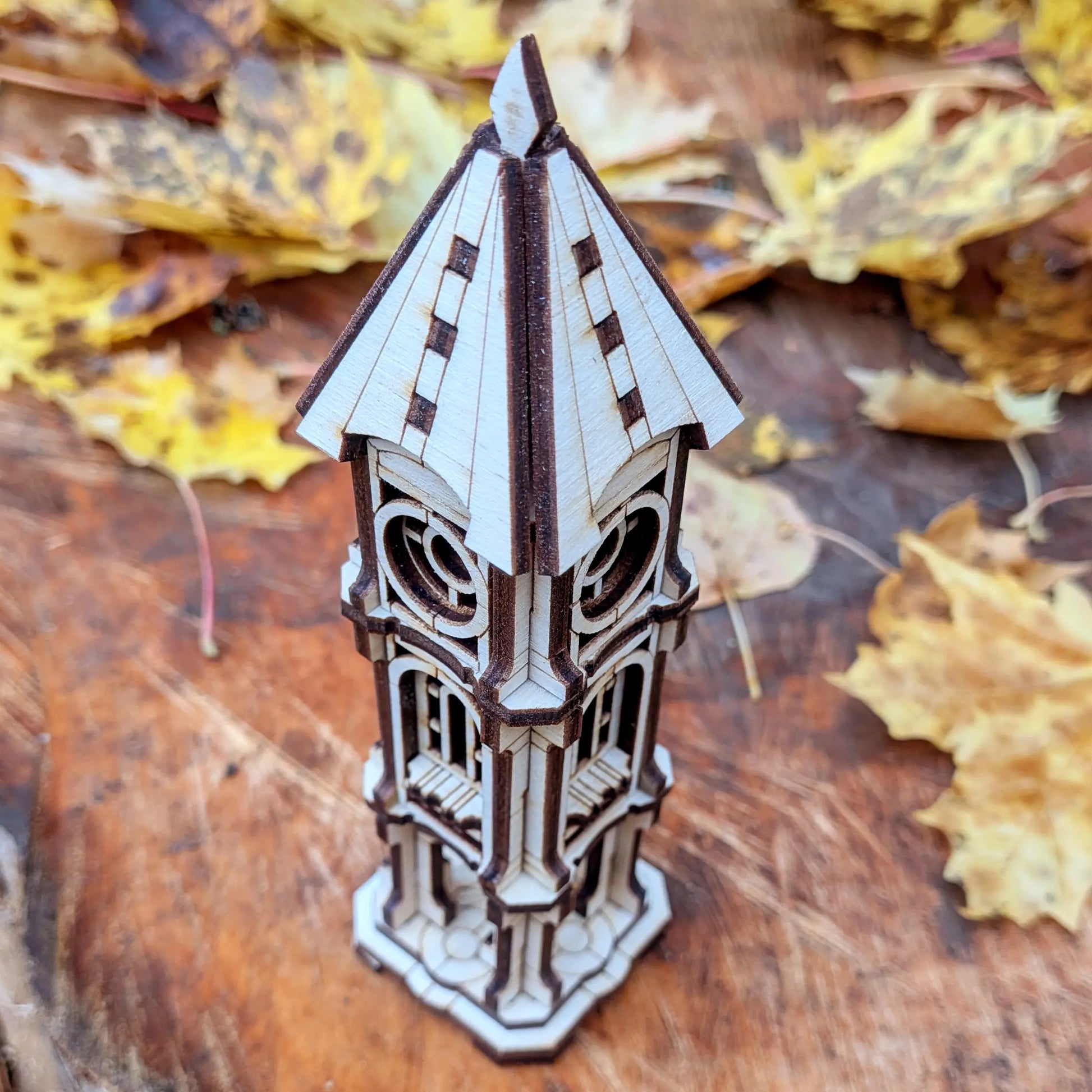 High-angle view of a miniature clock tower model made of laser cut plywood on a wood background with yellow autumn leaves