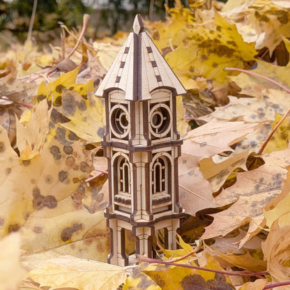 A miniature architectural clock tower model made of laser cut plywood beautifully nestled in a pile of yellow autumn leaves.