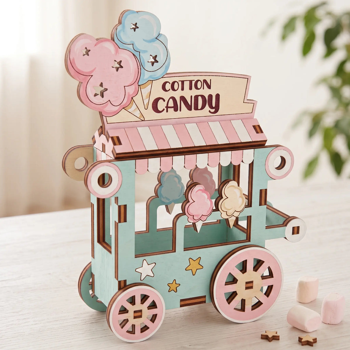 Painted pastel laser cut plywood cotton candy cart miniature with striped canopy and marshmallows on white wooden table.