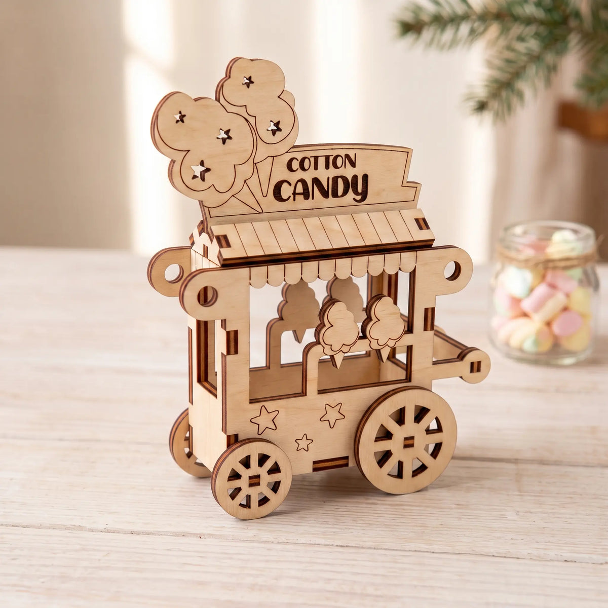 Laser cut plywood cotton candy cart model on rustic tabletop beside glass marshmallow jar and fresh evergreen pine branches.