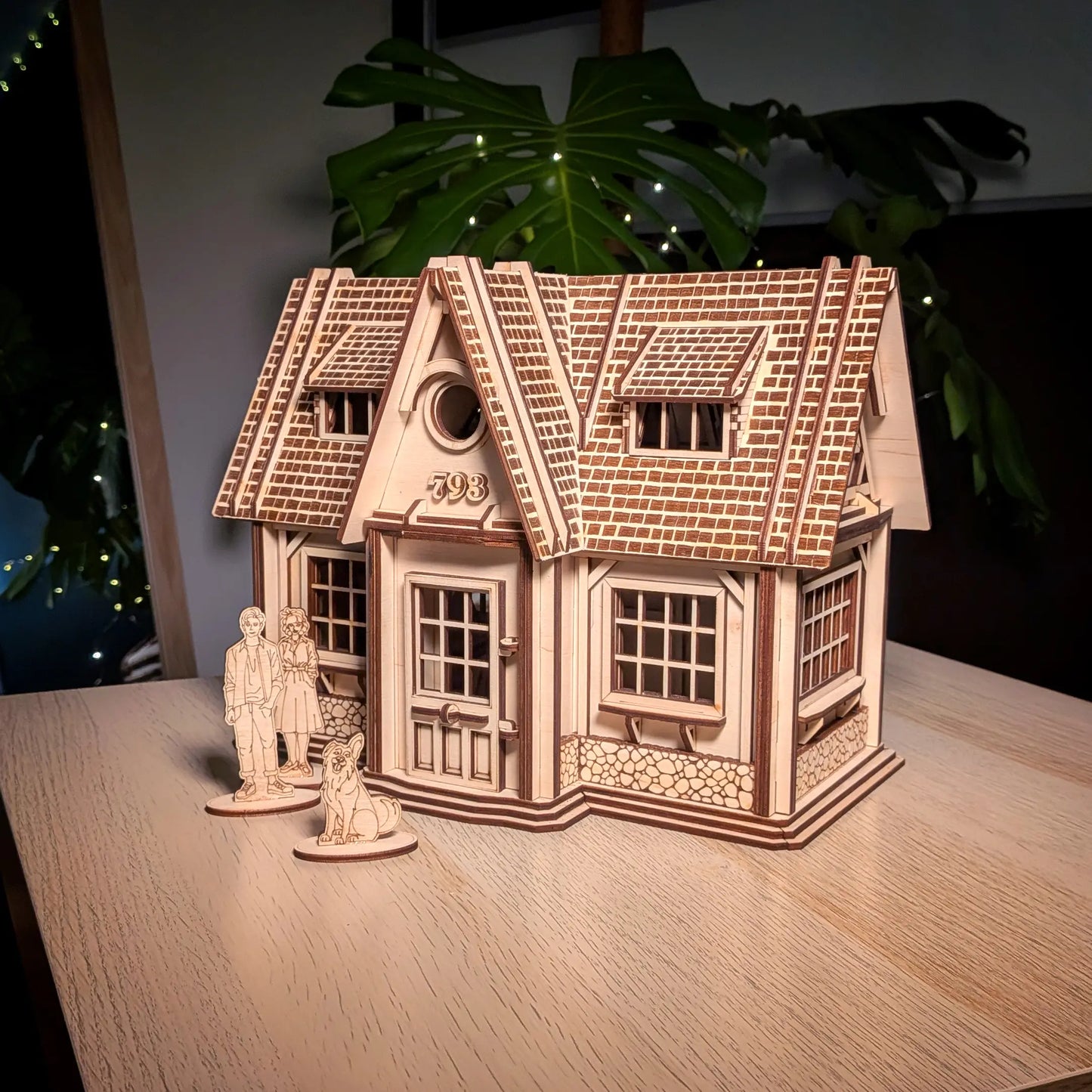 Finished laser cut plywood country house model on a wooden table with tiny family standees and a green leaf backdrop.