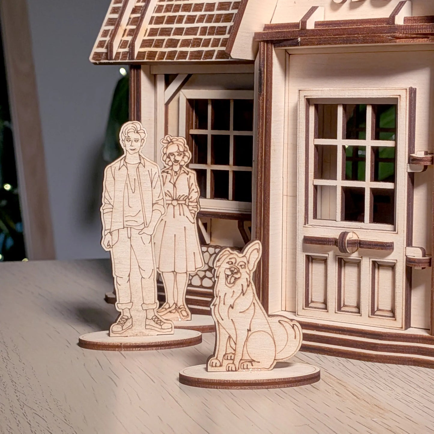 Miniature standee figurines of a man, woman, and dog placed on a wooden table before a laser cut plywood country house porch.