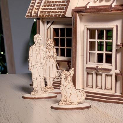Miniature standee figurines of a man, woman, and dog placed on a wooden table before a laser cut plywood country house porch.