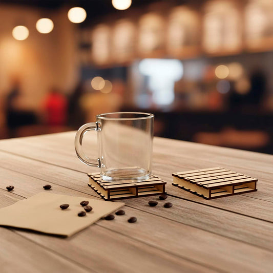 A clear glass mug sits on a laser cut wooden plywood mini-pallet coaster on a tabletop. A second empty pallet coaster rests nearby next to scattered coffee beans. The miniature pallet design features detailed slats and support beams