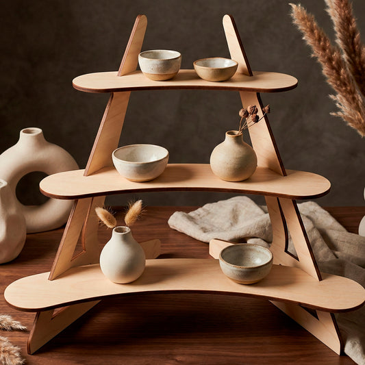 3-tier laser cut wooden plywood display stand made of smooth birch plywood, featuring curved shelves and a collapsible design. The retail riser is shown on a walnut table holding small handmade ceramics and dried pampas grass, illustrating a modern market display for craft fairs or boutiques