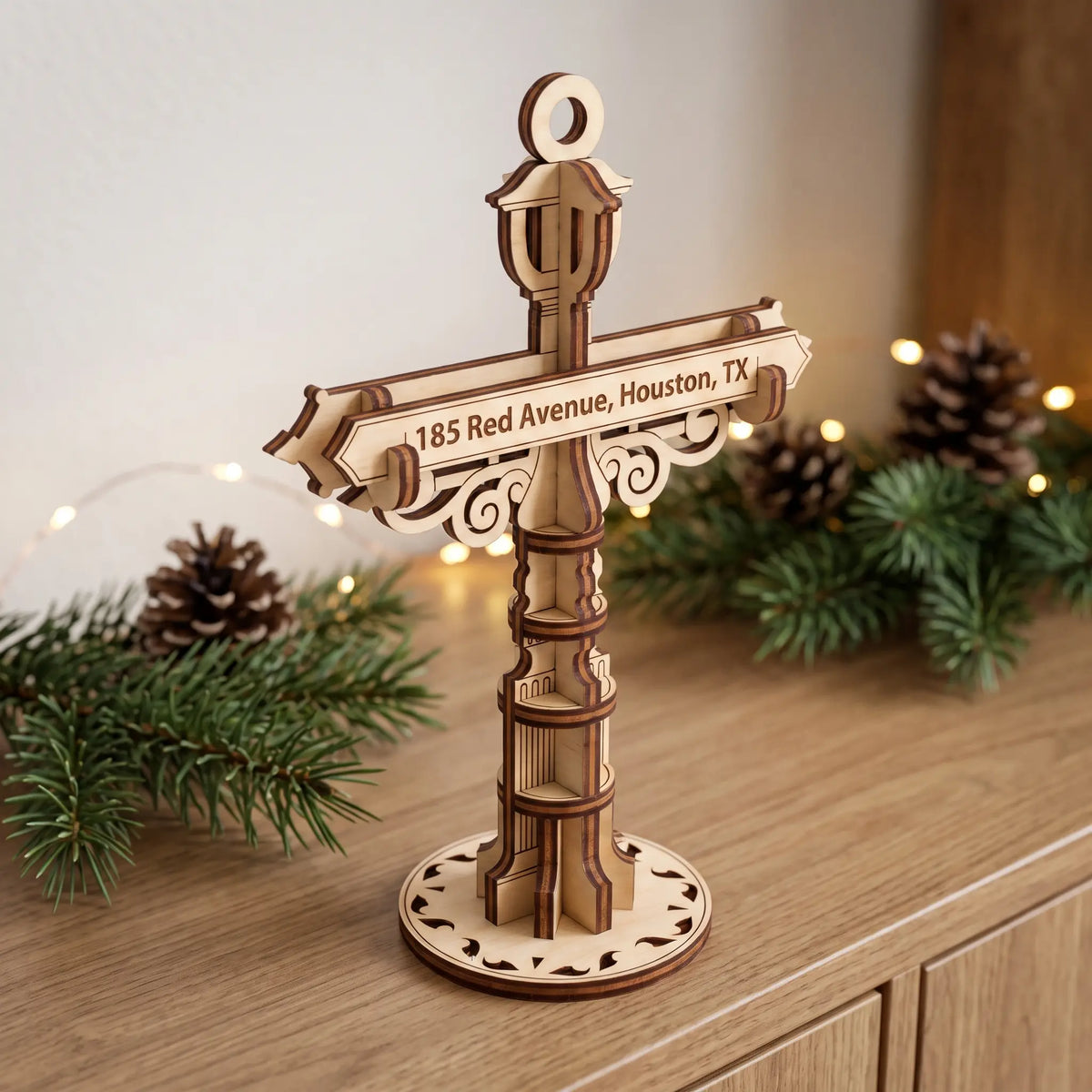 Laser cut plywood personalized street signpost ornament standing on wooden shelf beside pine branches and holiday lights.