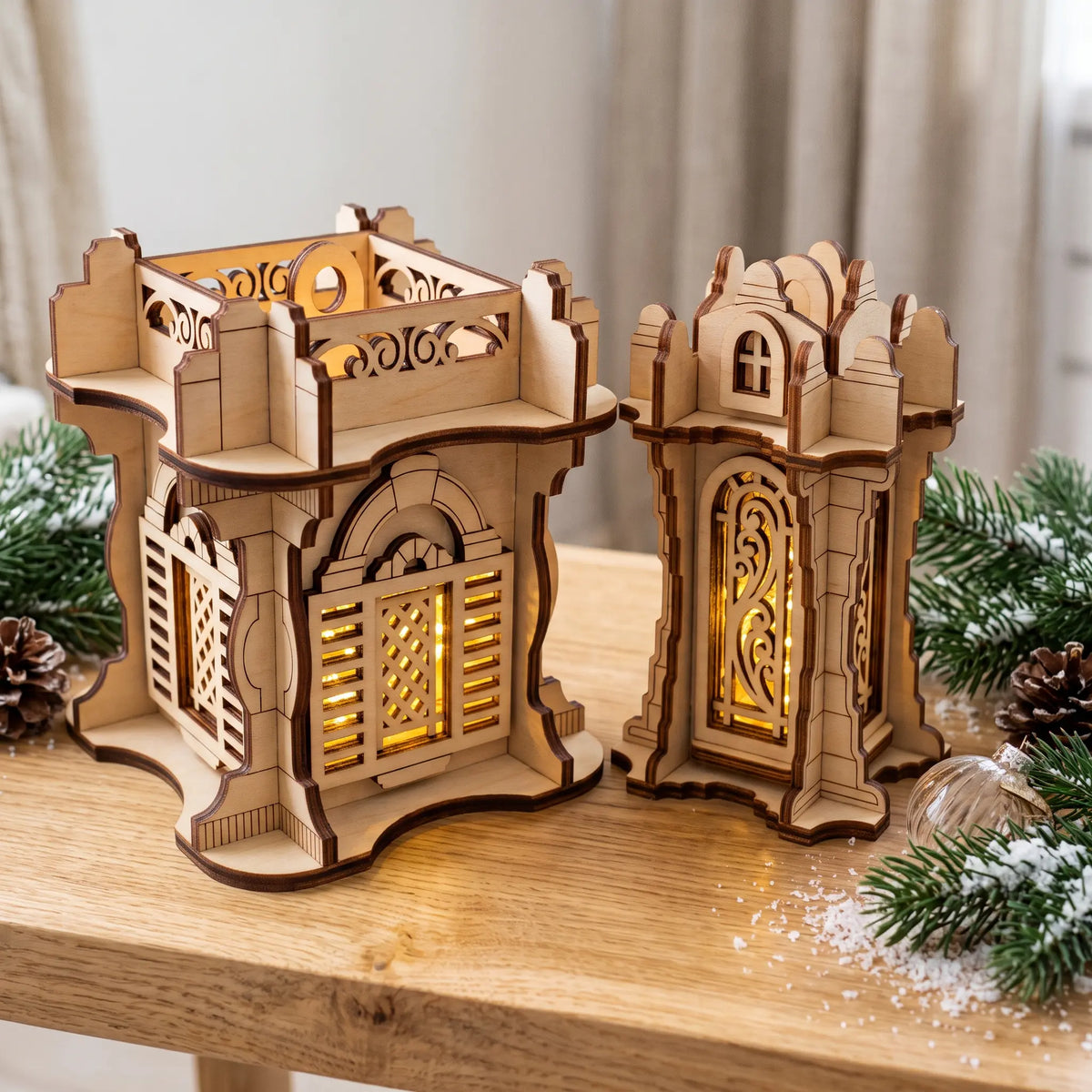 Two glowing laser cut plywood castle lanterns with carved lattice windows on wooden table next to pine branches and snow.