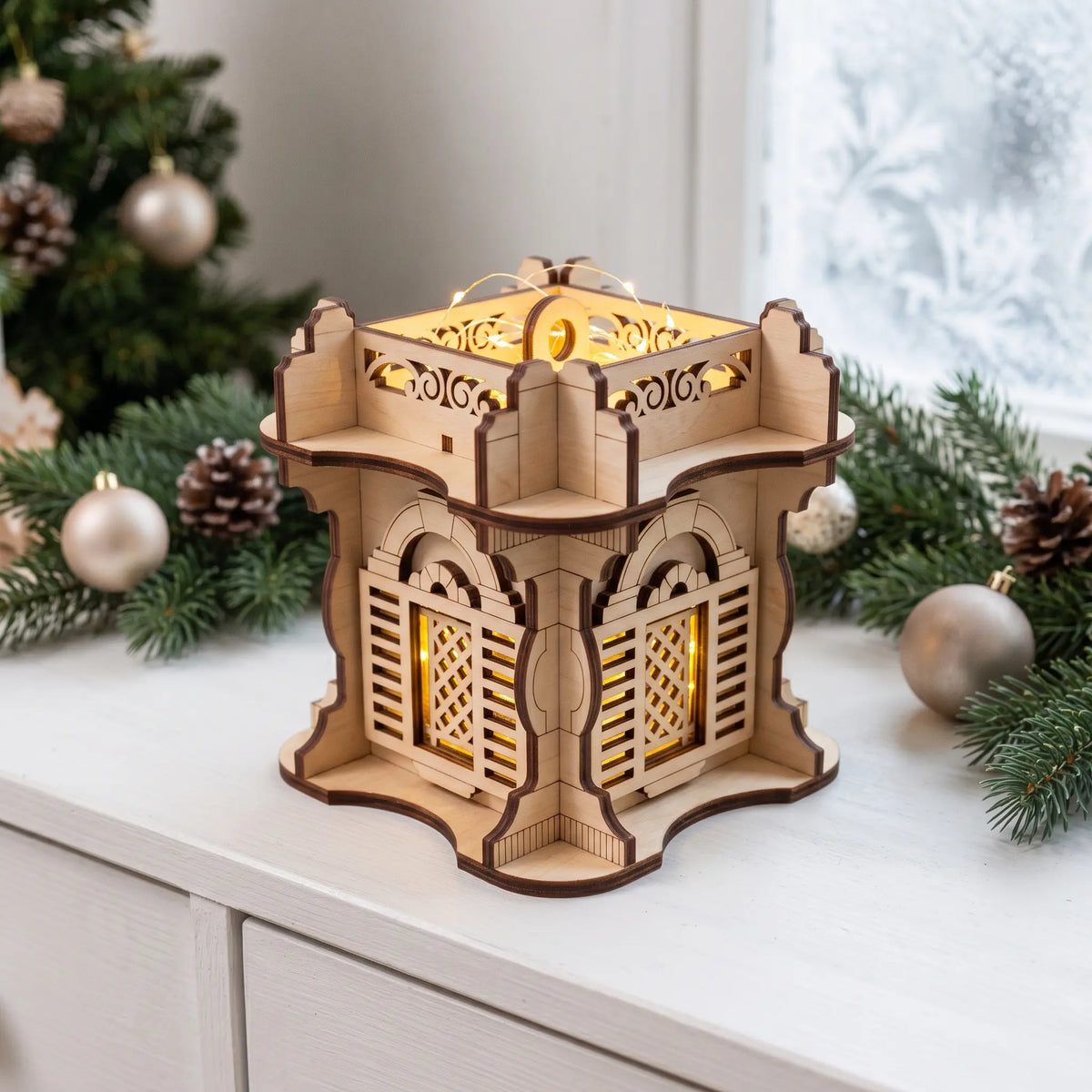 Warm glowing laser cut plywood square fortress lantern standing on white table beside evergreen branches and baubles.